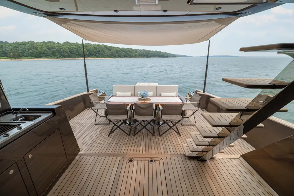 Aft cockpit under a deployed shade sail: dining set laid, transom sofa, wooded shoreline beyond.